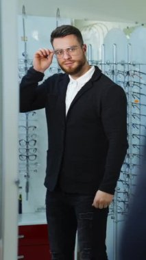 Optik dükkanında yeni gözlük deneyen bir adam. Yakışıklı adam yeni gözlük takıyor ve aynadaki yansımasına bakıp gülümsüyor. Göz giyimi konsepti. Dikey video
