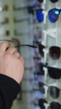 Modaya uygun güneş gözlükleri optikte duruyor. Dükkanda yeni gözlük deneyen bir adam. Alıcı optik mağazada gözlük seçiyor. Dikey video
