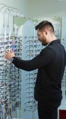 Optikteki yakışıklı adam. Yeni gözlük seçen genç bir adamın aynadaki yansıması. Erkek müşteri büyük aynanın yanında. Optometri. Dikey video