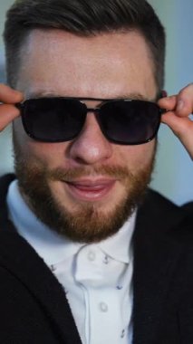 Dükkanda yeni siyah gözlüklü bir adamın yüz portresi. Güneş gözlüklü mutlu iş adamı kameraya bakıyor. Adam optik mağazada yeni gözlük seçiyor. Dikey video