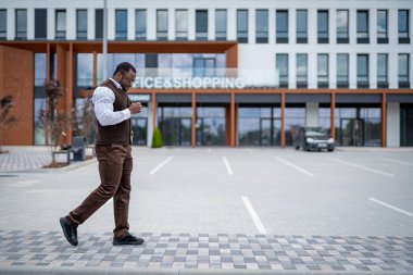 Ofis binasının önünde yürüyen adam. Kahverengi takım elbiseli ve beyaz gömlekli bir adam modern bir ofis binasının önündeki kaldırımda yürüyor..