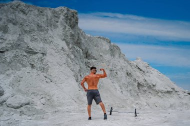 Beyaz kum tepeciğinin önünde kaslarını esneten adam. Gömleksiz adam beyaz kum tepeciğinin önünde kaslarını esnetir. Koyu renk şort giyiyor, siyah spor ayakkabılar, yerde halterli..