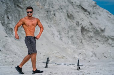 Taş ocağında kaslı adam halterin yanında poz veriyor. Kaslı bir adam elleri kalçasında poz veriyor, siyah şort ve güneş gözlüğü takıyor, taş ocağında bir halterin yanında..