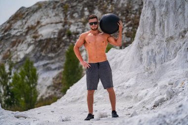 Maden ocağında Adam Tutma Balosu. Atletik şort ve güneş gözlüğü takan bir adam bir taş ocağında duruyor, omzunda bir ilaç topu tutuyor..