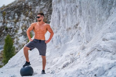 Taş ocağında tıp topu üzerinde duran adam. Kaslı bir adam arkasında beyaz bir kaya duvarıyla taş ocağında bir ilaç topunun üzerinde duruyor..