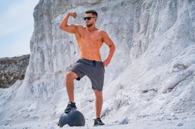 Taş ocağındaki kaslı adam. Bir adam taş ocağında ilaç topunun üzerinde dikilirken pazılarını esnetir..