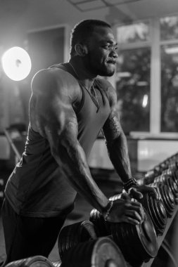 Spor salonunda Ağırlık Kaldıran Adam. Kaslı fiziği olan bir adam spor salonunda ağırlık kaldırır. Kolsuz tişört giyiyor ve antrenmanına odaklanmış durumda..