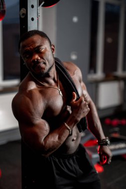 Spor salonunda kas yığını egzersizden sonra dinleniyor. Siyah şortlu kaslı bir adam ve omzuna havlu sarılmış bir adam spor salonunda duruyor. Antrenmandan sonra ara vermiş gibi görünüyor..