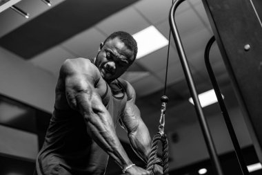 Spor salonundaki kablolu TV makinesinde çalışan adam. Bir adam spor salonundaki kablolu TV makinesinde çalışıyor. Ağırlığı kaldırmaya, pazılarını esnetmeye odaklanmış. Görüntü siyah beyaz..