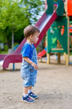 Çocuk çocuk oyun sahasında duruyor. Tulum giymiş genç bir çocuk oyun parkında duruyor, arka plandaki kırmızı kaydırağa bakıyor..
