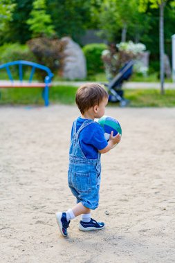 Toddler Park 'ta topla oynuyor. Genç bir çocuk elinde renkli bir topla parkta kumlu bir patikada yürür. Mavi bir tulum ve mavi bir tişört giyiyor..