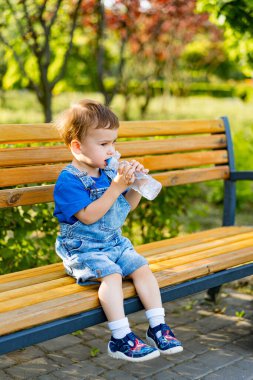 Park 'taki Bench' te su içen çocuk. Genç bir çocuk bir bankta oturuyor, tulum ve mavi bir gömlek giyiyor, bir şişeden su içerken..