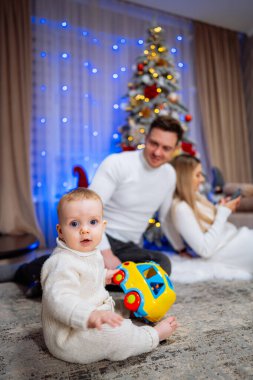 Bebek, Noel Ağacı 'nın yanında oyuncak arabayla oynuyor. Beyaz kazaklı bir bebek oyuncak bir arabayla oynarken ailesi arka planda süslü bir Noel ağacının yanında oturuyor..