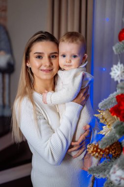 Anne Bebeği Tutuyor Noel Ağacı 'nın yanında. Anne kameraya gülümsüyor bebeği bir Noel ağacının yanında tutuyor, bebek başka yere bakıyor..