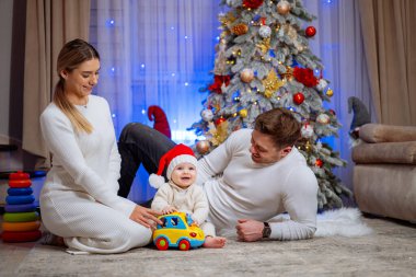 Aile, Noel Ağacı 'nın yanında Bebekle oynuyor. Üç kişilik bir aile Noel fotoğrafı için poz veriyor, anne babalar süslenmiş bir Noel ağacının önünde bebekleriyle etkileşime geçiyor..
