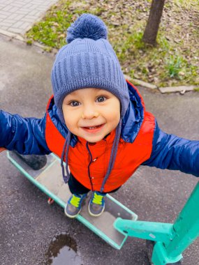 Mavi şapkalı çocuk dışarıda scooter sürüyor. Mavi şapkalı ve kırmızı ceketli genç bir çocuk kaldırım yolunda scooter sürerken gülümsüyor..