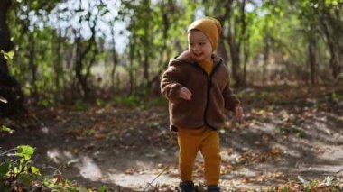 Parkta güzel beyaz bir bebek koşuyor. Sonbahar doğasında yürüyen şapkalı ve ceketli gülümseyen bir çocuk..