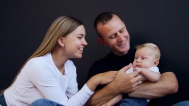 Babasının elinde oturan sevimli sarışın bebek. Anne sevimli oğluyla oynuyor. Siyah arkaplan.