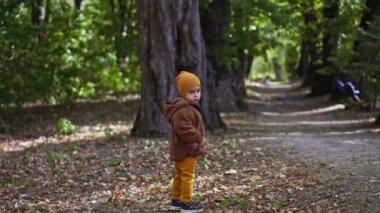 Sakin, ciddi, sıcak kıyafetler giyen erkek bebek parkta duruyor. Çocuk sonunda gülümsedi ve uzaklaştı..
