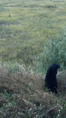 Çimenlerdeki karanlık ve ürkütücü bir figürün üst görüntüsü. Siyah mantolu, kapüşonlu bir hayalet sahada tek başına duruyor. Cadılar Bayramı zamanı. Dikey video