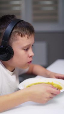 Çocuk dijital tablete bakarken evde yemek yiyor. Kulaklıklı çocuk mutfakta masada oturuyor ve yemek yiyor. Gadget 'lar ve çocuklar. Dikey video