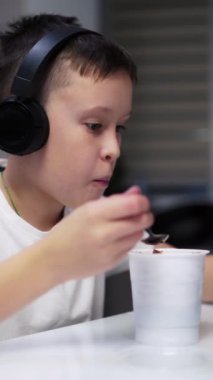 Çocuk mutfakta yemek yiyor. Siyah kulaklıklı çocuk evde masada yemek yerken bir tabletle oynuyor. Dikey video