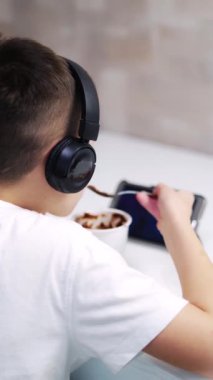 Çocuk video izleyip evde yemek yiyor. Mutfaktaki masada oturan ve tablete bakan kulaklıklı bir çocuğun arka görüntüsü. Dikey video