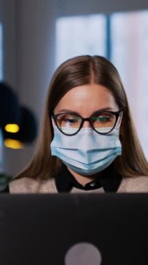 Maskeli genç bayan çalışan bilgisayarda çalışıyor. Gözlüklü kadın bilgisayarını kapatıyor ve doğrudan kameraya bakıyor. Dikey video