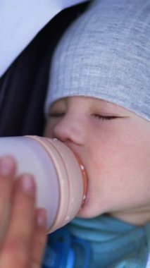 Anne küçük oğlunu bir şişeyle besliyor. Güzel çocuk bebek arabasında yatarak yemek yiyor ve uykuya dalıyor. Kapatın. Dikey video