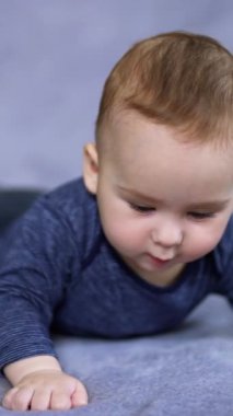 Mavi elbiseli sevimli beyaz çocuk ayaklarını sallayarak göbeğinde yatıyor. Meraklı çocuk, kaşlarını yavaşça kaldıran bir ilgiyle yukarı bakıyor. Dikey video