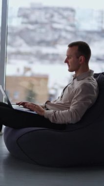 Rahat bir odada dizüstü bilgisayarda çalışan serbest çalışan bir erkek işadamı. Panoramik pencerelerin ve bulanık kışın şehir manzarasının arka planında koltukta oturan bir adam. Dikey video