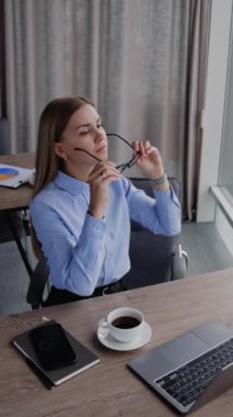 Ofis çalışanı gözlük takıp çalışmaya başlıyor. Kadın panoramik pencerenin yanındaki masada dizüstü bilgisayarda çalışıyor. Yüksek açı. Dikey video