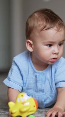 Mavi elbiseli şirin erkek bebek yerde dört ayak üstünde duruyor. Sevimli çocuk önünde bir oyuncak seçmeye çalışıyor. Arkaplan bulanık. Dikey video