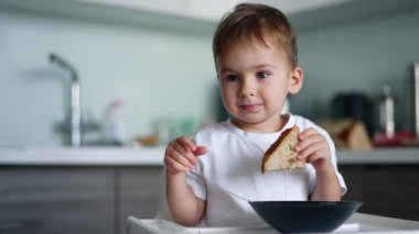 Sevimli erkek bebek elinde bir parça ekmek tutarak yüksek masada oturuyor. Sevimli çocuk ileriye bakıyor dikkatlice oturuyor..