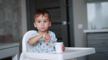 Yoğurttan hoşlanan sevimli erkek çocuk. Bebek mutfakta oturup plastik bardaktan süt ürünleri yiyor. Bulanık arkaplan.