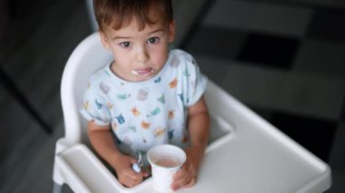 Küçük çocuk elinde yoğurtla beyaz sandalyede oturuyor. Bebek, biraz süt ürünleri alıp yemek için kaşık kullanıyor. Üst Manzara. Bulanık arkaplan.