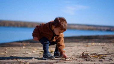 Kahverengi ceket ve kot pantolon giyen beyaz bir çocuk rıhtımda yürüyor. Çocuk iki taş seçip nehre atıyor..