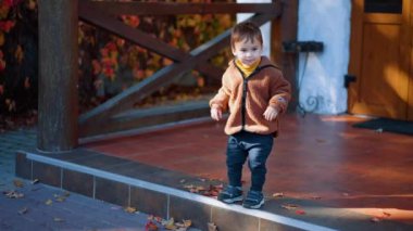 Kahverengi ceketli şirin erkek bebek verandada duruyor. Gülen çocuk merdivenden mutlu bir şekilde atlıyor..