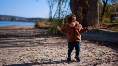 Sevimli küçük çocuk nehir kıyısında bir şey aramak için yürüyor. Güneşli bir sonbahar gününde doğada sevimli bir çocuk..