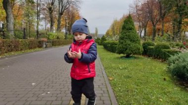 Parkta yürürken ceketli ve şapkalı sevimli bir oğlan. Çocuk kameraya doğru geliyor kolundaki su damlalarını gösteriyor..