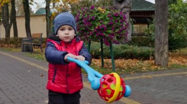 Elinde oyuncak olan beyaz bir erkek çocuk. Yeni yürümeye başlayan çocuk sıcak kıyafetler giyiyor sonbaharda parkta yürüyüş yapıyor..
