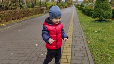Şapkalı ve ceketli sevimli çocuk parkta. Aktif çocuk mutlu bir şekilde atlıyor ve gülümsüyor..