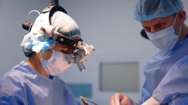 Genç kadın beyin cerrahı ameliyatta. Doktor prosedürde cihaz gözlüğü kullanıyor. Erkek asistan cerraha yardım eder.