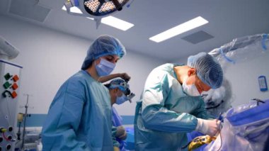 Metal aletlerle hastanın üzerine eğilmiş profesyonel beyin cerrahı. Personele yardım etmek yanında duruyor.