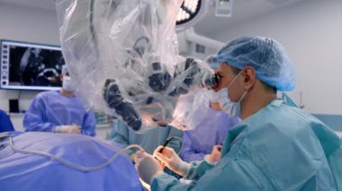 Odaklanmış beyin cerrahı, mikroskoba dikkatlice bakan hastayı ameliyat eder. Maskeli doktor meslektaşlarıyla konuşuyor..