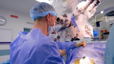 Ameliyat odasındaki mikroskobun yanında oturan bir erkek cerrahın dikiz görüntüsü. Doktor metal forseps ve cerrahi iplik kullanıyor..