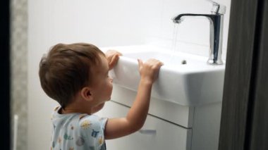 Banyoda lavabonun yanında durup zıplayan küçük mutlu çocuk. Yürüyen çocuk çeşmeden akan suyun tadını çıkarıyor..