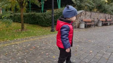 Kırmızı ceketli küçük çocuk kameradan kaçıyor. Sevimli çocuk sonbahar günü parkın önünden geçiyor..