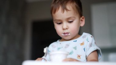 Dudaklarında süt ürünleri olan sevimli bir erkek çocuk. Bebek fincandan yoğurt yiyor. Düşük açı. Bulanık arkaplan.