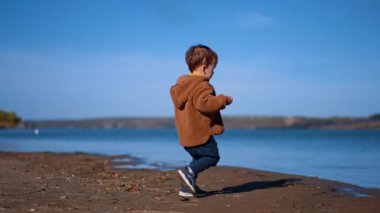 Kahverengi ceketli ve pantolonlu çocuk nehre koşuyor. Çocuk iki eliyle de taş atıyor. Sonbaharda güneşli bir gün.